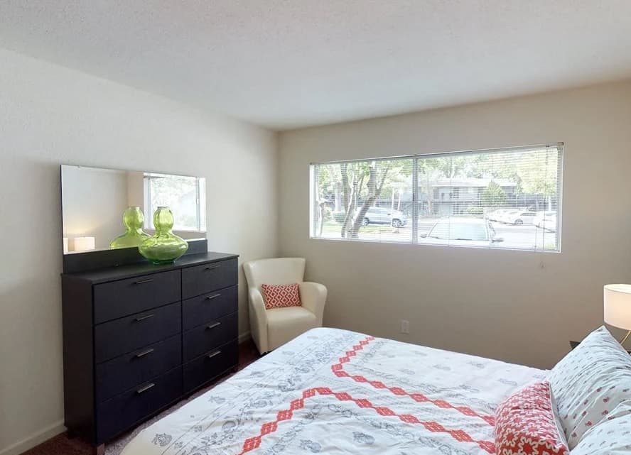 A bedroom at Westpark Plaza with a large window, white walls, a bed with a patterned white and red comforter, a black dresser with a mirror and green glass vases, and a white armchair with a red pillow.