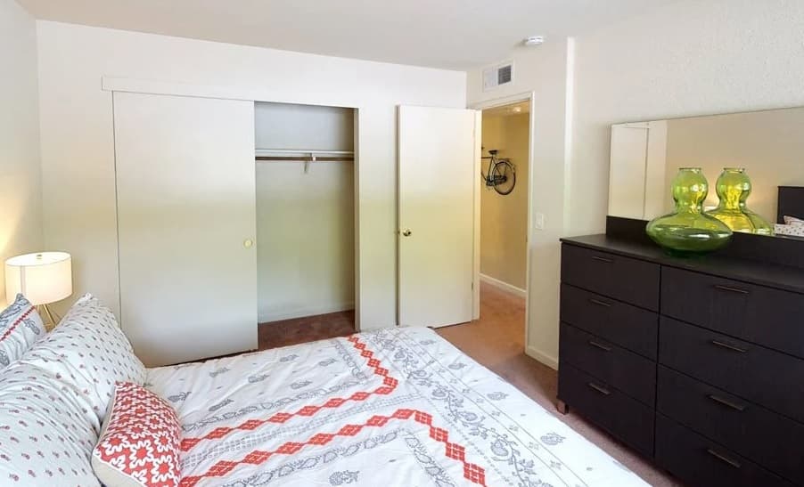 A tidy Westpark Plaza bedroom with a bed covered in patterned white bedding, a dark dresser with green glass vases, a bedside lamp, and an open closet with sliding doors. Light neutral walls and carpet flooring complete the look.