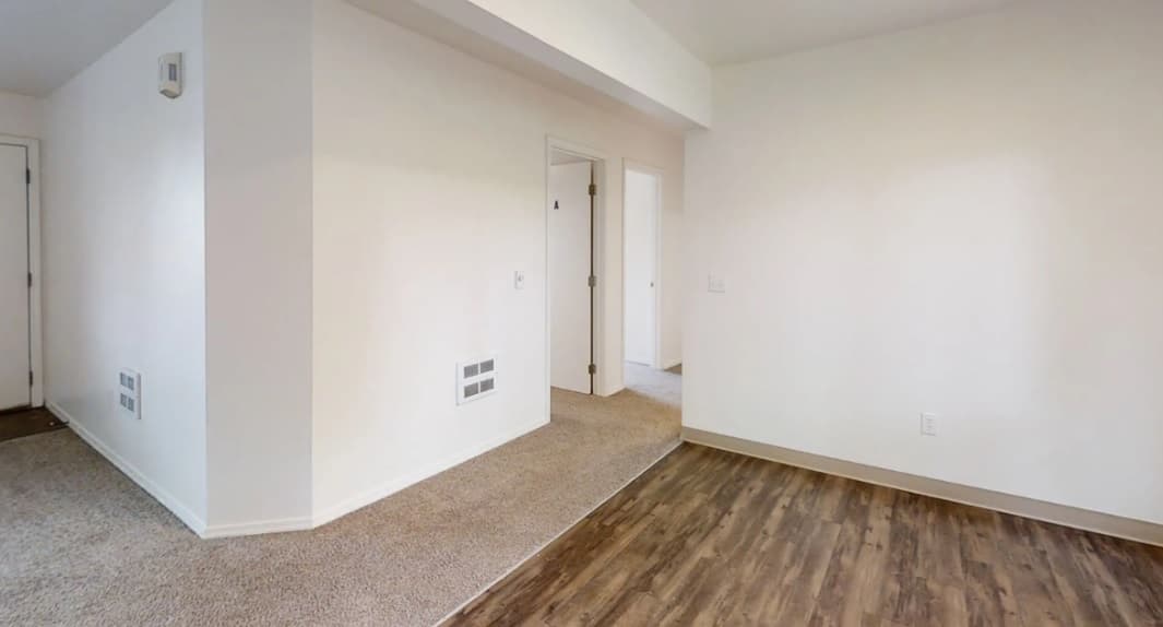 A room at Willow Place with white walls, wood-look flooring on the right, and carpeted flooring on the left. An open doorway leads to another room, with a hallway visible in the background.