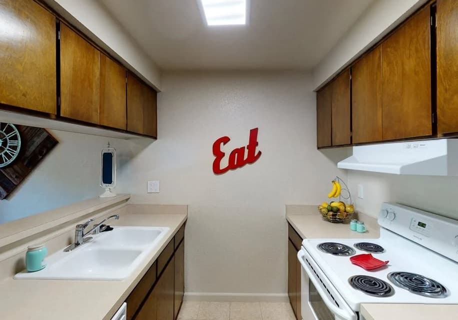 A small galley kitchen in Westpark Plaza features wooden cabinets, white countertops, a white stove and sink, fruit on the counter, and a red wall sign that says Eat above the back wall.