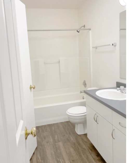 A clean, modern bathroom at Willow Place features white walls, a bathtub-shower combo, toilet, white vanity with gray countertop, sink, mirror, and wood-look flooring. The space is well-lit and simple in design.
