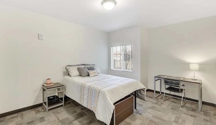 Minimalist bedroom in Wolf Run Vintage Reno style with a neatly made single bed, side table, desk with lamp, chair, and window letting in natural light. Neutral tones and simple decor create a clean, uncluttered space.