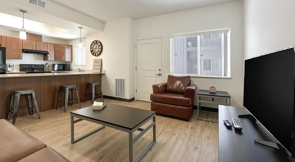 Modern apartment living room with wood floors, brown leather chair, and Wolf Run Vintage Reno touches. The open kitchen features barstools, pendant lights, stainless steel appliances, and a large window that brightens the inviting space.