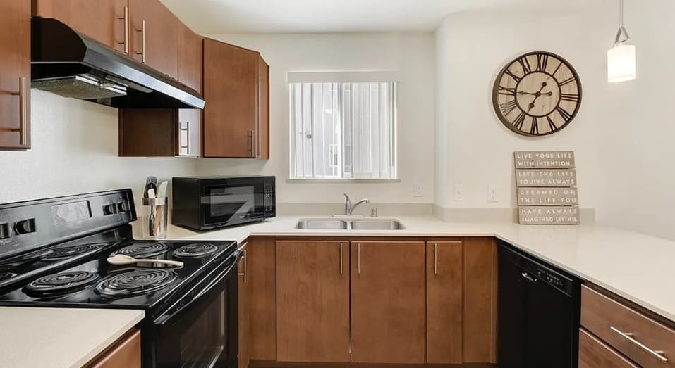 Modern kitchen at Wolf Run Student Housing Apartments Reno with brown cabinets, black appliances, microwave, double sink under a window, wall-mounted clock, and decorative sign on a light countertop. Bright, clean, and well-organized space.