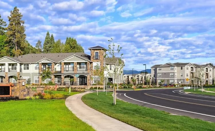 A modern suburban apartment complex in Easton with well-kept lawns, young trees, and a curved road under a partly cloudy blue sky. Multiple two-story buildings are surrounded by greenery and a clean sidewalk.