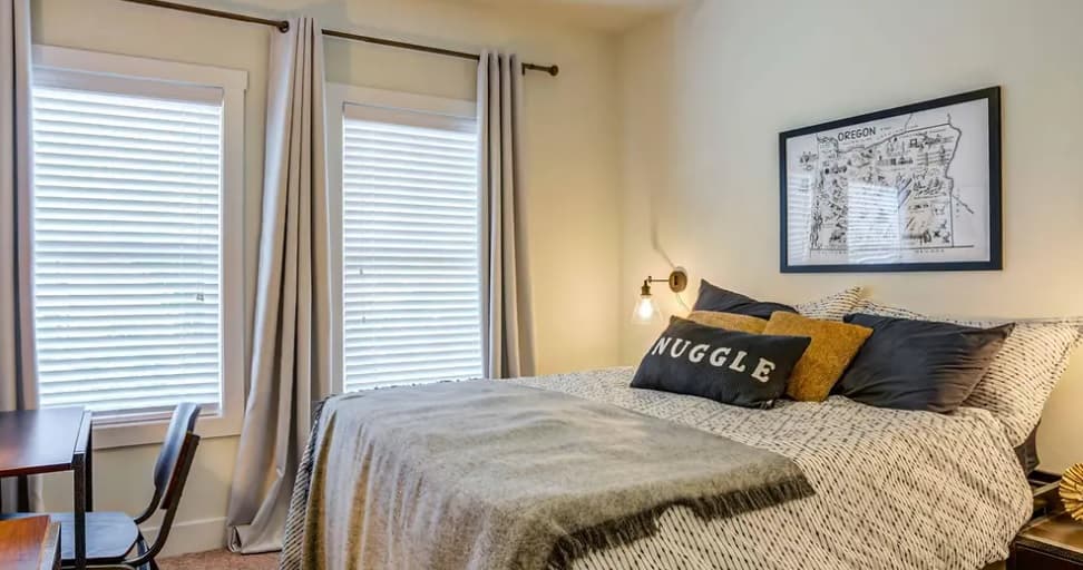 A cozy bedroom in Yugo Corvallis with a bed covered in neutral bedding, accent pillows (one reads SNUGGLE), a gray throw, two large windows with white blinds and curtains, a framed map on the wall, and a small desk with a chair.