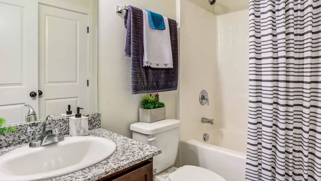 A modern bathroom at Yugo Corvallis with a granite countertop, sink, and faucet on the left, a toilet in the center, a bathtub with a striped shower curtain, and towels above the toilet. A small plant decorates the toilet tank.