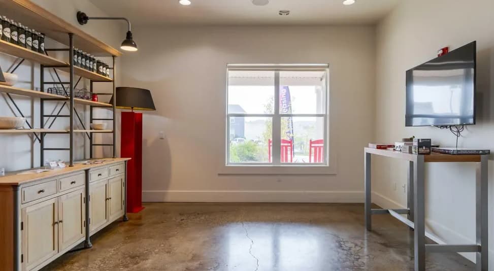 A modern student accommodation at Pullman Hills featuring concrete floors, a large window, wall-mounted TV, tall bookshelf with jars and decor, white sideboard, red floor lamp, and a table with board games for fun evenings.