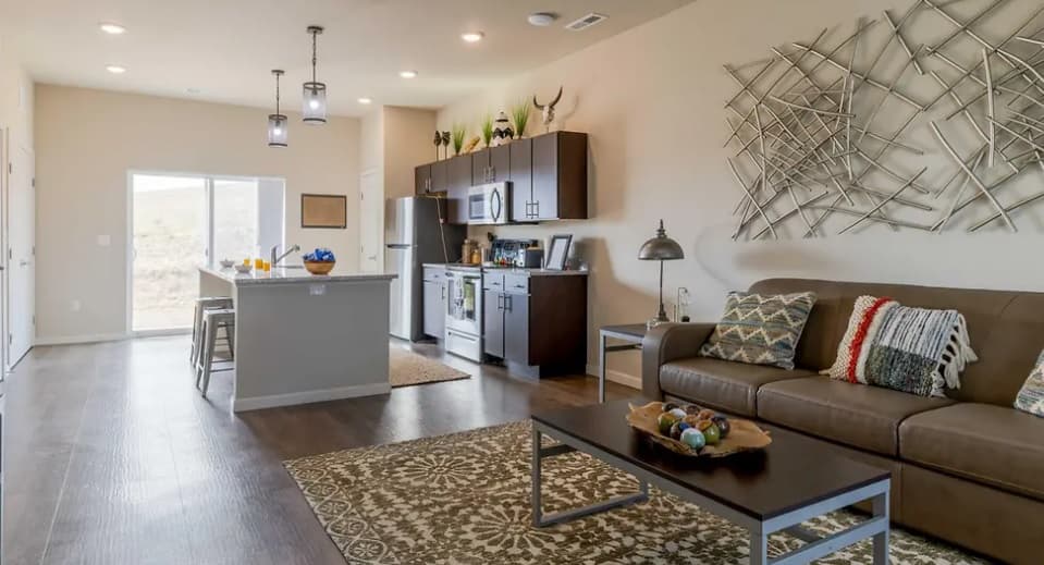 Modern open-concept kitchen and living room at Pullman Hills with brown cabinets, stainless steel appliances, a tan sofa with colorful pillows, patterned rugs, and abstract wall art. Large windows let in natural light.