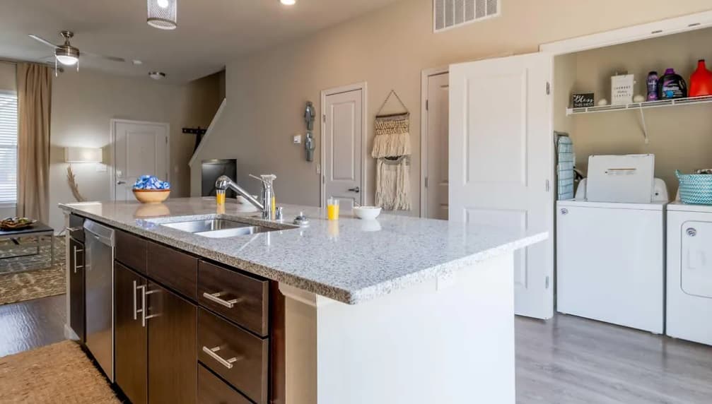 Modern kitchen with granite island, sink, and dark wood cabinets. In the background, a laundry area with washer, dryer, and shelves is visible through open double doors. Warm lighting brightens this inviting Pullman Hills Yugo residence.