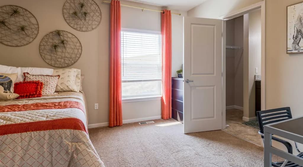 A cozy Yugo Pullman Hills bedroom with a neatly made bed, decorative pillows, wall art, and bright orange curtains. A dresser sits nearby, with an open closet door and a partial view of a desk and chair completing the inviting space.