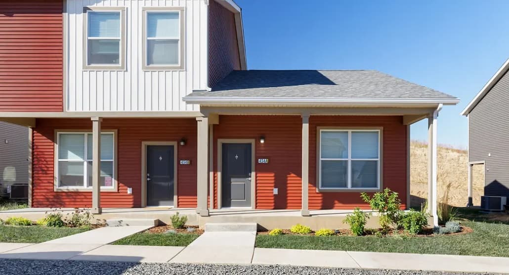 A modern duplex home at Pullman Hills with red siding and white trim, two separate entrances, small front porches, neatly landscaped lawns, and a bright blue sky in the background.