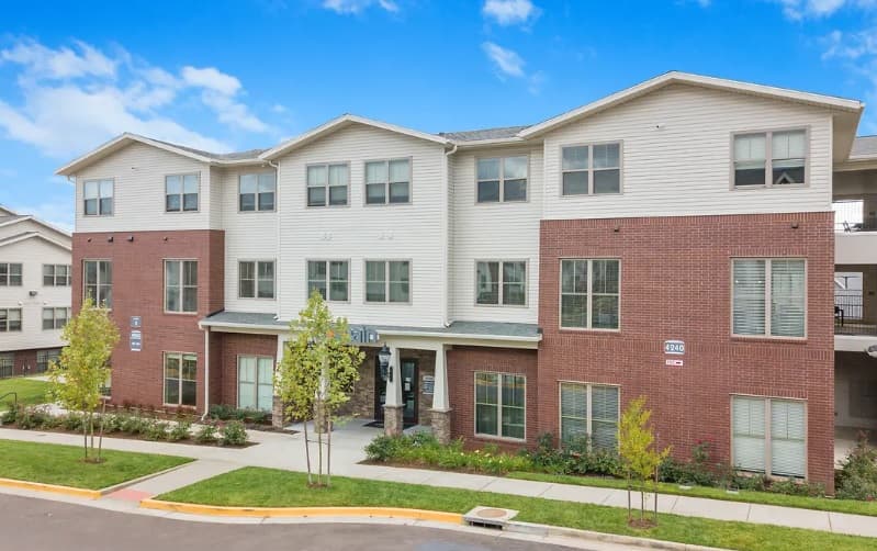 A modern three-story apartment building in Corvallis, perfect for student accommodation, with red brick on the lower level, white siding above, large windows, a covered entrance, and small trees along the sidewalk under a bright blue sky.
