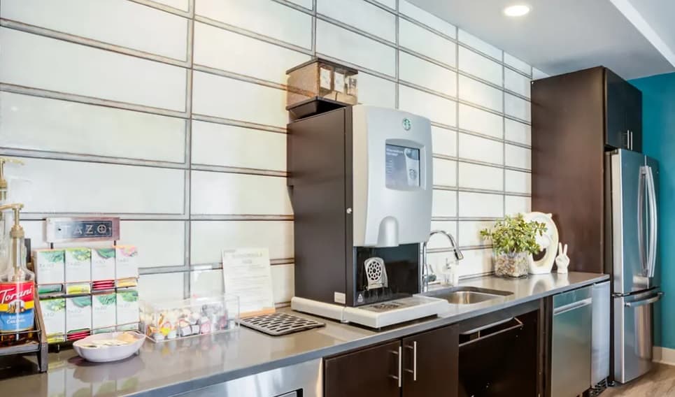 A modern kitchen counter at Yugo Corvallis features a coffee and water machine, a sink, assorted tea and coffee supplies, a plant, a refrigerator, and cabinetry, set against a tiled white-and-gray wall in this student housing space.