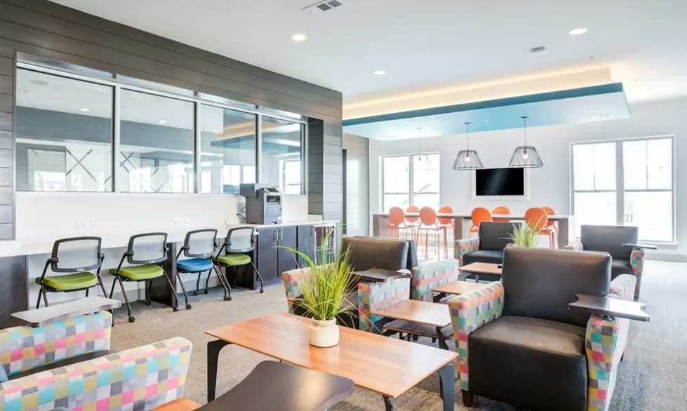 Modern lounge in Yugo Corvallis with patterned armchairs, small wooden tables with plants, black chairs at a counter, orange barstools, large windows, and a flat-screen TV. A bright, airy space perfect for relaxing Corvallis student housing.
