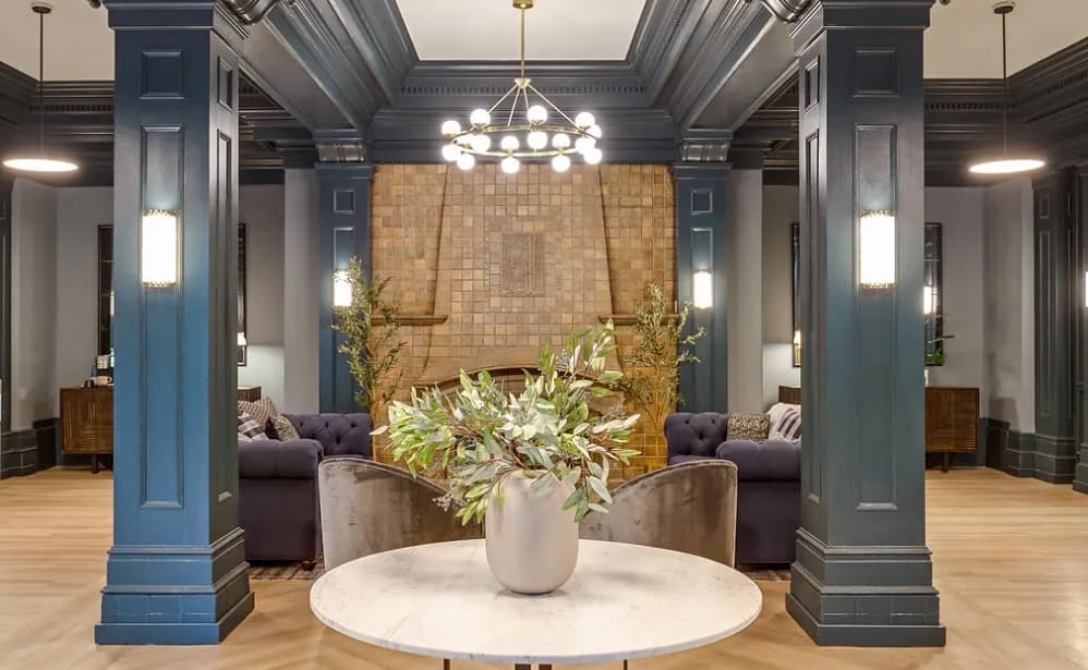 A stylish lobby at Yugo Seattle with dark blue columns, a round marble table topped with a vase of greenery, and a sitting area featuring tufted sofas and a tiled fireplace in the background offers Wilsonian elegance.