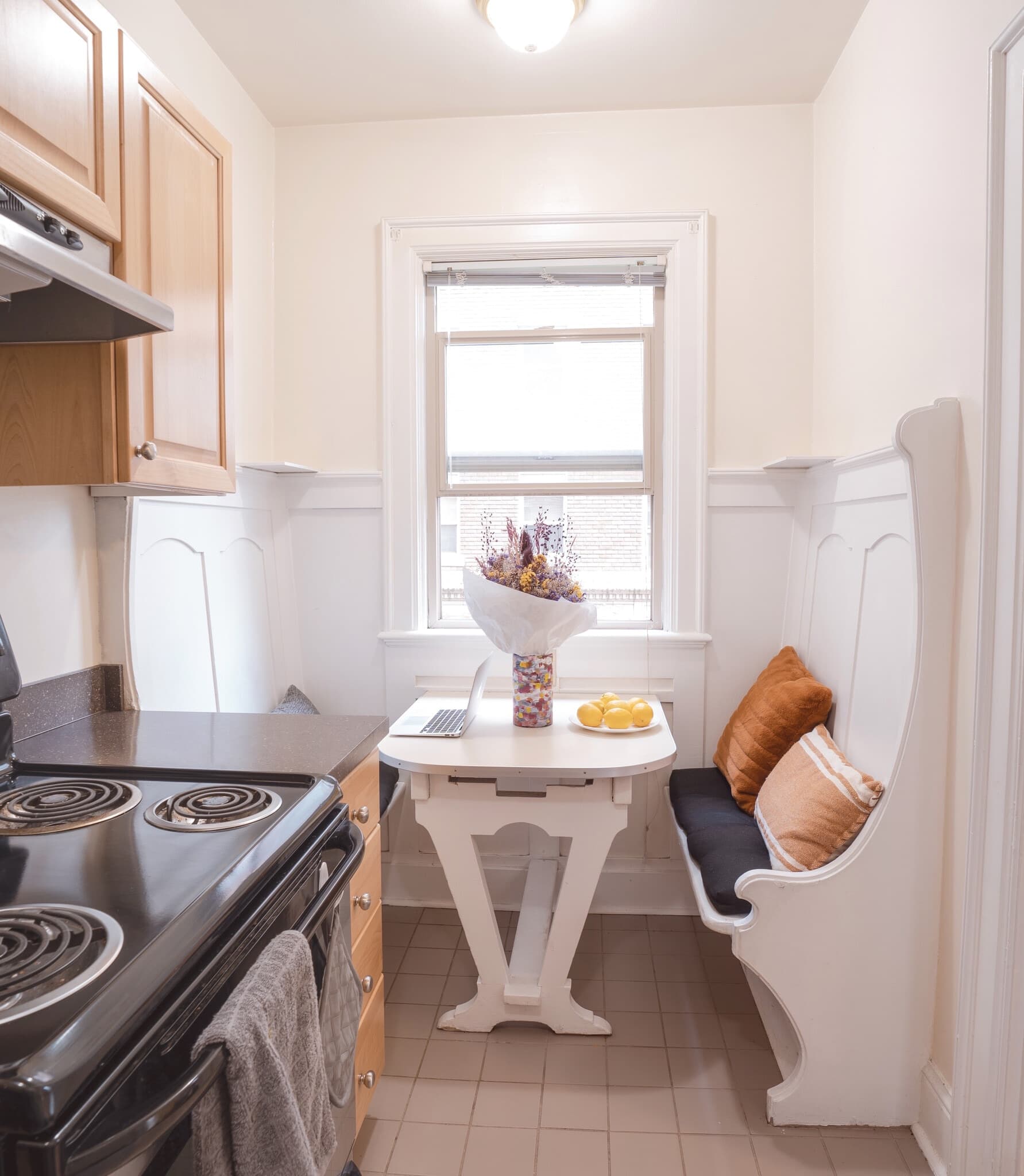 A small, bright kitchen nook in Yugo Seattle with a white bench and table, two brown cushions, a vase of flowers, lemons on a plate, and a laptop. Nestled by the window near the stove and wooden cabinets, it's ideal for Wilsonian-inspired mornings.