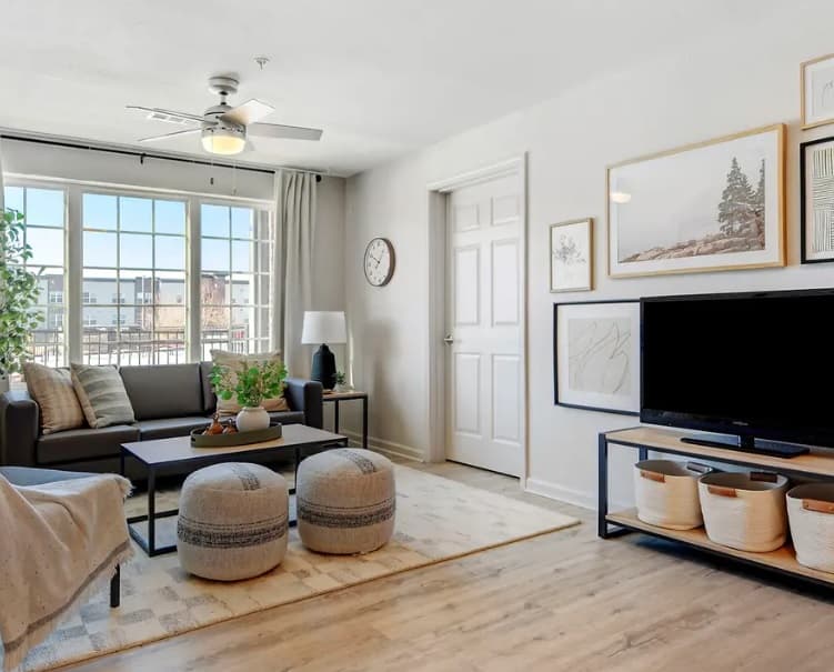 Bright living room at Yugo Fort Collins, featuring large windows, a gray sofa, two round poufs, coffee table, TV on a stand with baskets, wall art, houseplants, clock, and light wood flooring. Neutral tones and cozy decor create a welcoming space.