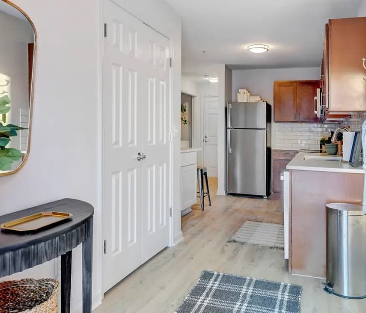 Modern kitchen at Yugo Fort Collins with wood cabinets, stainless steel refrigerator, white tile backsplash, light wood floors, a black side table with a mirror, and double white door closet. A small rug lies in front of the kitchen area.