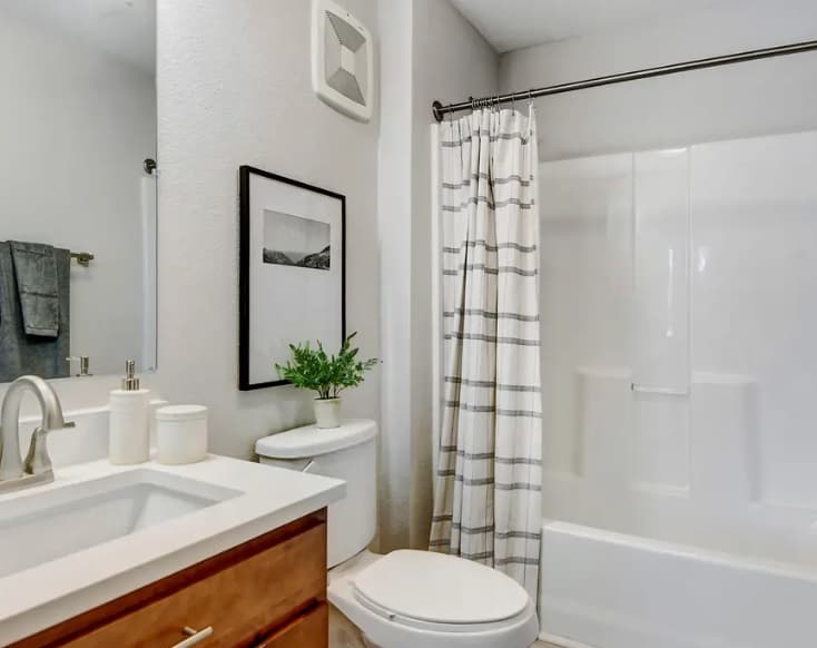 A modern bathroom at Yugo Fort Collins features a white sink, wooden vanity, toilet, potted plant, wall art, and a bathtub with a striped shower curtain. Towels hang on the wall, and the neutral color scheme creates a clean look.