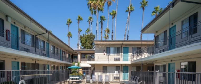 A two-story motel with teal doors and railings, surrounding a sunny outdoor pool area with lounge chairs and palm trees, evokes the relaxed vibe of SDSU weekends beneath a clear blue sky.