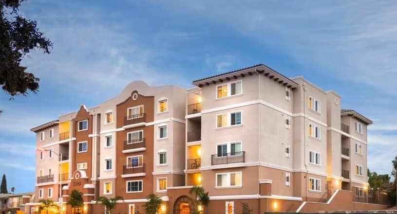 A four-story student housing building with light brown and beige walls, multiple windows, balconies, and decorative architectural details, photographed at dusk under a blue sky.