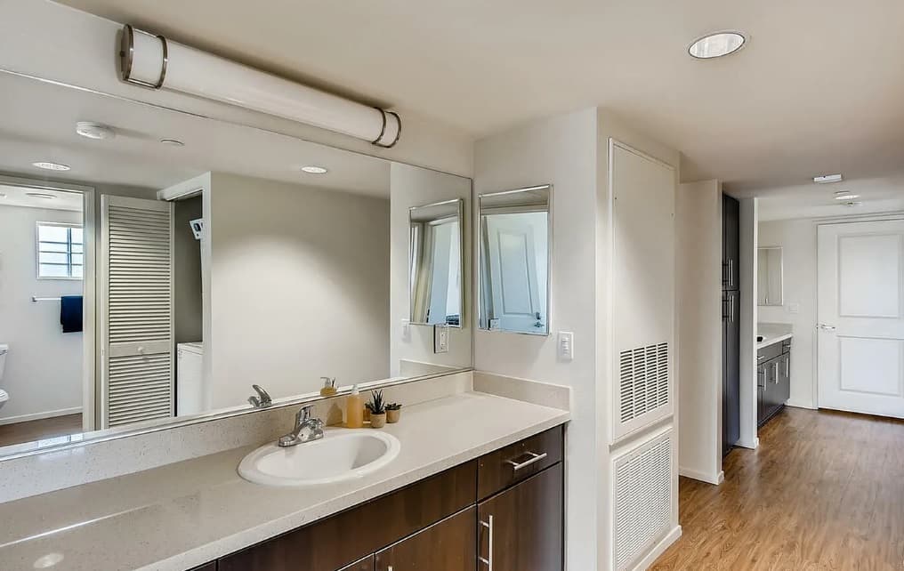 Modern bathroom in Zuma Student Housing with a large mirror above a sink and wooden vanity, light fixture on the ceiling, and hallway leading to another room with wood flooring. Reflections reveal extra cabinets and bathroom space.