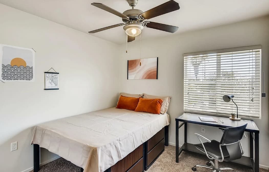 A neatly made bed with orange pillows sits beside a window with blinds in this Zuma Student Housing room. A desk with a chair, lamp, and notebook is nearby. The space features light walls, a ceiling fan, carpeted floor, and minimal wall art.