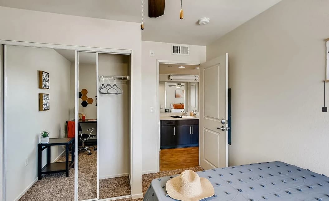 A tidy bedroom in Zuma student housing with a bed, beige hat on top, mirrored closet doors, and an open door to a modern bathroom. A small workspace with a chair and decor is reflected in the closet, perfect for student accommodation.