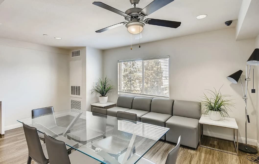 Modern living and dining area in Zuma Student Housing with a glass table, gray chairs, gray sectional sofa, two potted plants, a floor lamp, ceiling fan, and a window letting in natural light.