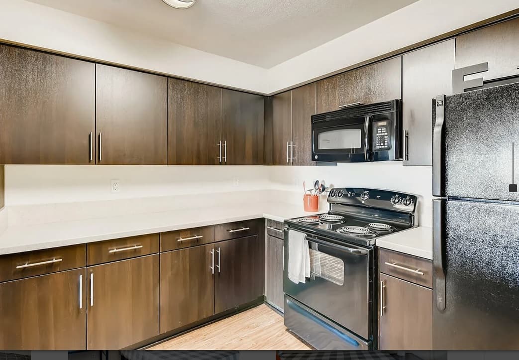Modern kitchen in Zuma Student Housing with dark wood cabinets, white countertops, black appliances—including stove, microwave, and refrigerator—and a few utensils and a pot on the stove, offering a clean and organized appearance.