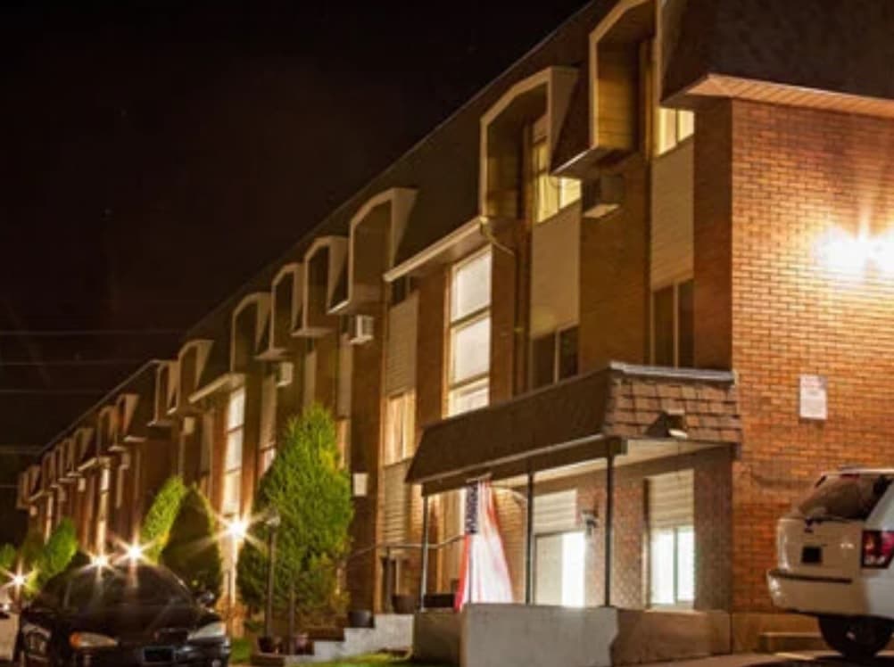 A row of brick Bridgerland Apartments Logan buildings at night, warmly lit by exterior lights, with parked cars and small trees lining the walkway.