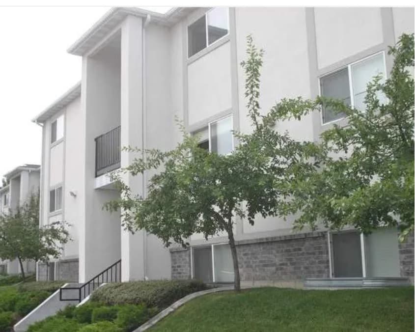 Three-story Oakridge Student Apartments Logan building with light-colored walls and large windows, surrounded by green trees and shrubs. Steps and a small black railing lead to a side entrance.