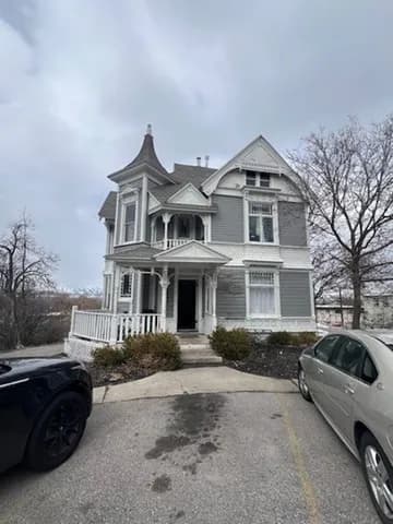 A large Victorian-style gray and white house with a turret, porch, and ornate trim—reminiscent of the charm found at UV Apartments Logan—stands among bare trees and parked cars on a cloudy day.