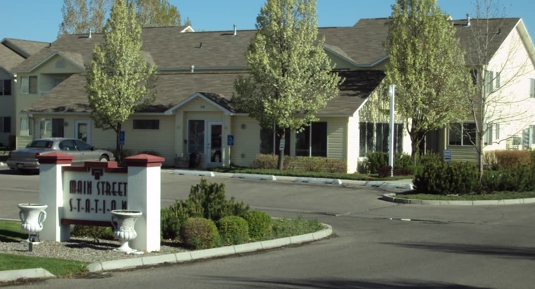 A one-story, light-colored building with a brown roof and several trees in front. A red and white sign at the entrance reads Main Street Station Apartments. A few parked cars are visible in the lot.