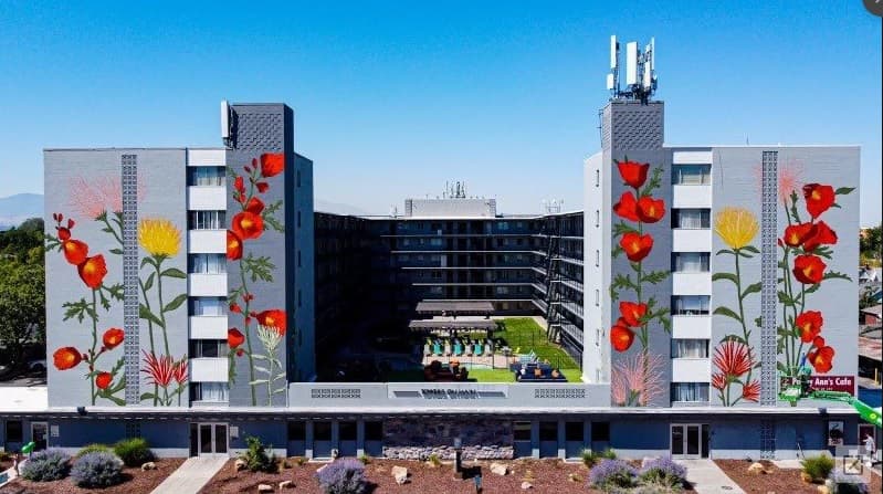 A modern apartment building with two tall towers featuring large, colorful floral murals. Towers on Main Salt Lake City frame a central courtyard with a pool. The scene is bright and clear, with landscaping in front of the building.