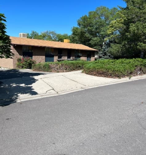 A single-story brown brick building with a tan roof sits at 2122 S Nevada Ave, Provo, set behind shrubs and a concrete walkway, bordered by trees under a clear blue sky.
