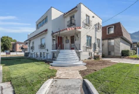 La Casa Provo is a two-story white stucco apartment building with red doors, a small staircase leading to the entrance, and a neatly trimmed lawn under a clear blue sky.