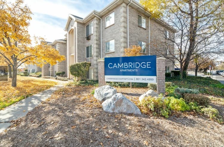A beige brick apartment building with autumn trees in front and a large blue sign that reads Cambridge Apartments Provo, featuring contact information, all surrounded by attractive landscaping and rocks.