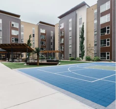 Outdoor view of Cedar Heights Rexburg, a modern apartment complex with tall beige and brown buildings, a covered patio area with seating, landscaped greenery, and a blue basketball court in the foreground.