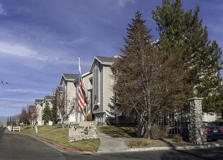 A row of beige apartment buildings with peaked roofs, an American flag, trees with bare branches, and a sign reading Country Sun and Country Springs Orem sits beside the street under a blue sky with some clouds.