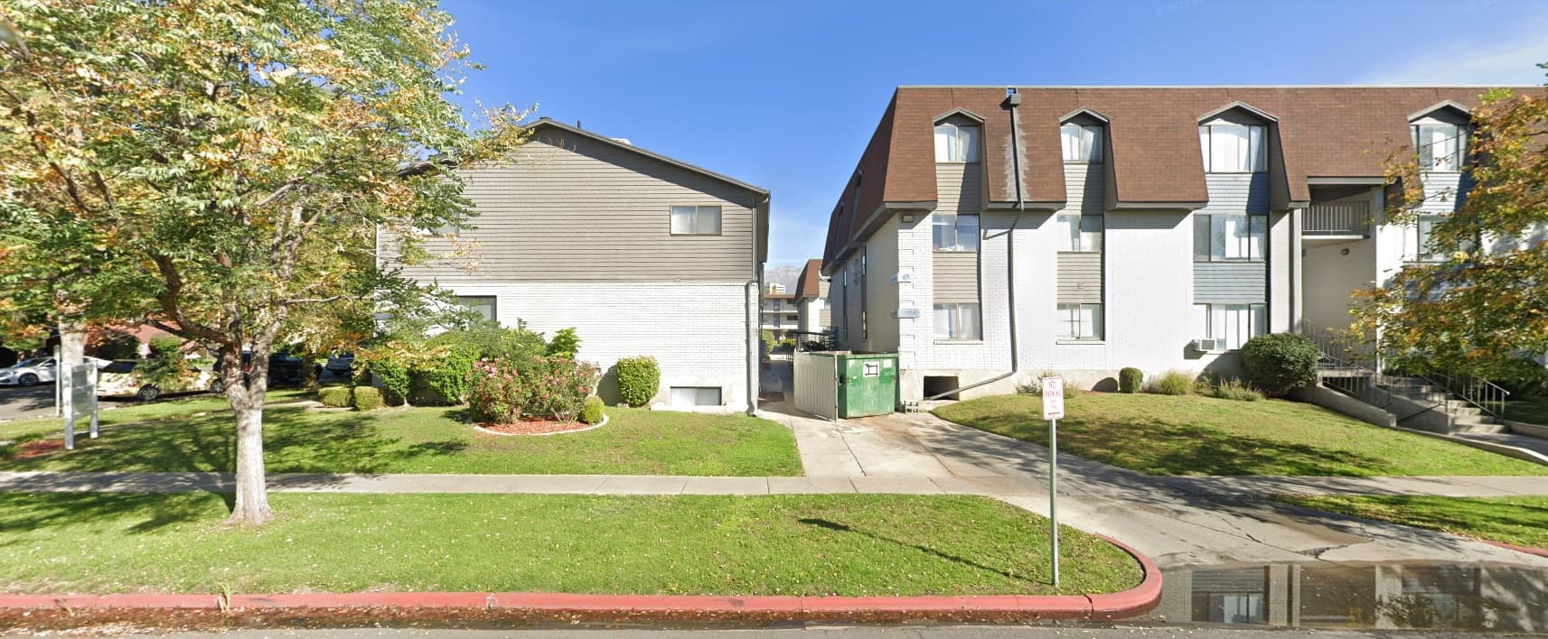 Two residential buildings with light-colored exteriors, part of Green Gables Provo, are separated by a driveway with a green dumpster. The area features green lawns, trees, shrubs, and a sidewalk alongside a red-painted curb.