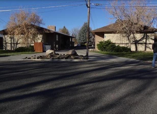A quiet suburban street with two single-story houses, leafless trees, and power lines—just steps from Homestead Villas Rexburg. Rocks line the driveway as a person walks away on the right; shadows stretch across this peaceful neighborhood scene.