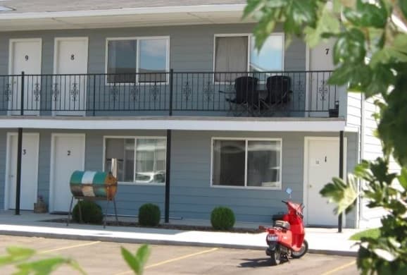 A small motel with gray siding and black railings, showing two floors of rooms numbered 1 to 9, resembles the cozy charm of Lexington Apartments Rexburg. A red scooter is parked out front, with a barbecue grill near the lower rooms and green leaves framing the image.