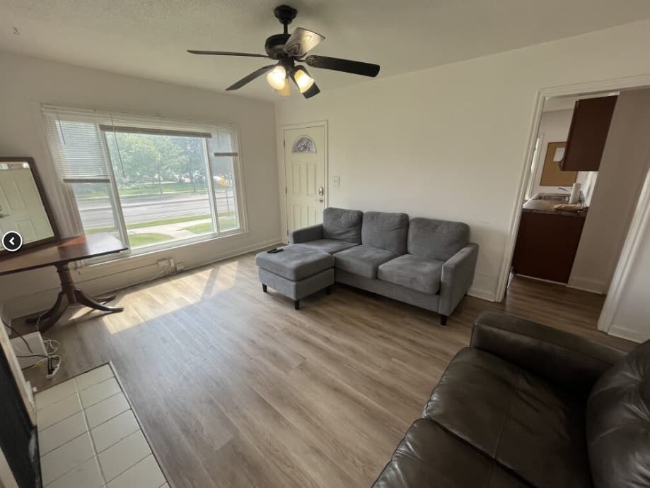 A bright living room at Lookout House Provo features a large window, gray sectional sofa, brown leather couch, wood-look flooring, ceiling fan with lights, and a small dining table near the window. A doorway leads to the kitchen area.