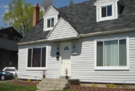 A white, single-story Marshall Duplex in Provo with an attic, large front windows, a small porch with steps, and a neatly trimmed lawn bordered by bricks under a clear blue sky.