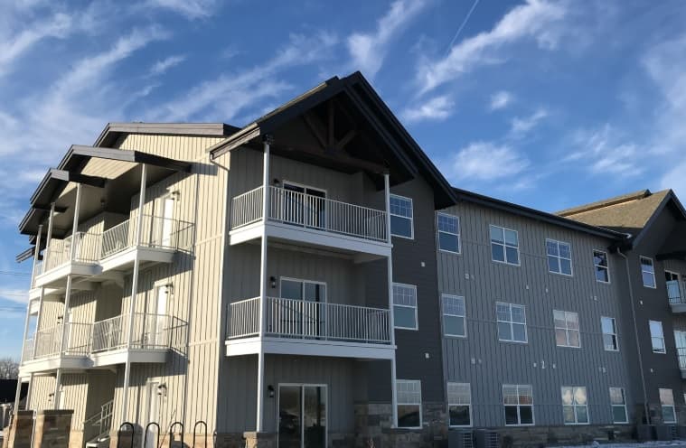 Three-story modern apartment building at Mountain Shores Rexburg with light gray siding, white railings on balconies, and large windows, pictured under a blue sky with scattered clouds.