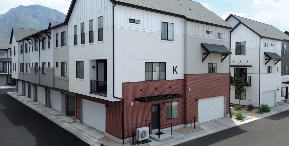 A modern three-story townhouse building at Oakpointe Provo features white siding, red brick on the lower level, black trim, and multiple garages. The building is labeled “K” and sits on a clean, empty street with mountains in the background.