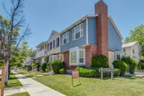 Two-story suburban house with blue-gray siding, brick chimney, white trim, and shutters, on a sunny day. Located near Promenade Provo, it features a green lawn, shrubs, sidewalk in front, and a For Sale sign in the yard.