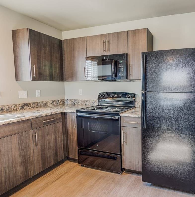 A modern Rocky Ridge Rexburg kitchen with wood cabinets, granite countertops, black appliances such as a refrigerator, electric stove, and microwave, and stylish light wood flooring.